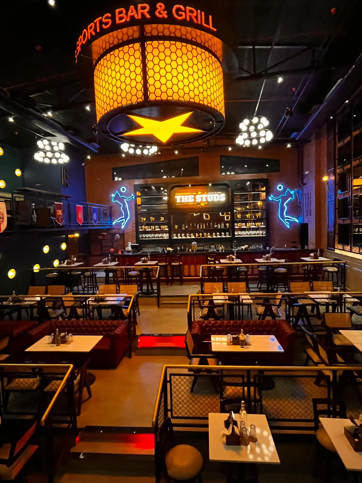 The main seating floor of The Studs in Ranchi, featuring industrial-style decor, tufted booths, and neon sports player lights on the brick wall.