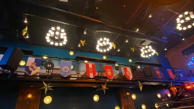 The upper deck of The Studs sports bar in Ranchi, showcasing various sports team flags hanging from the railing and unique circular light fixtures.